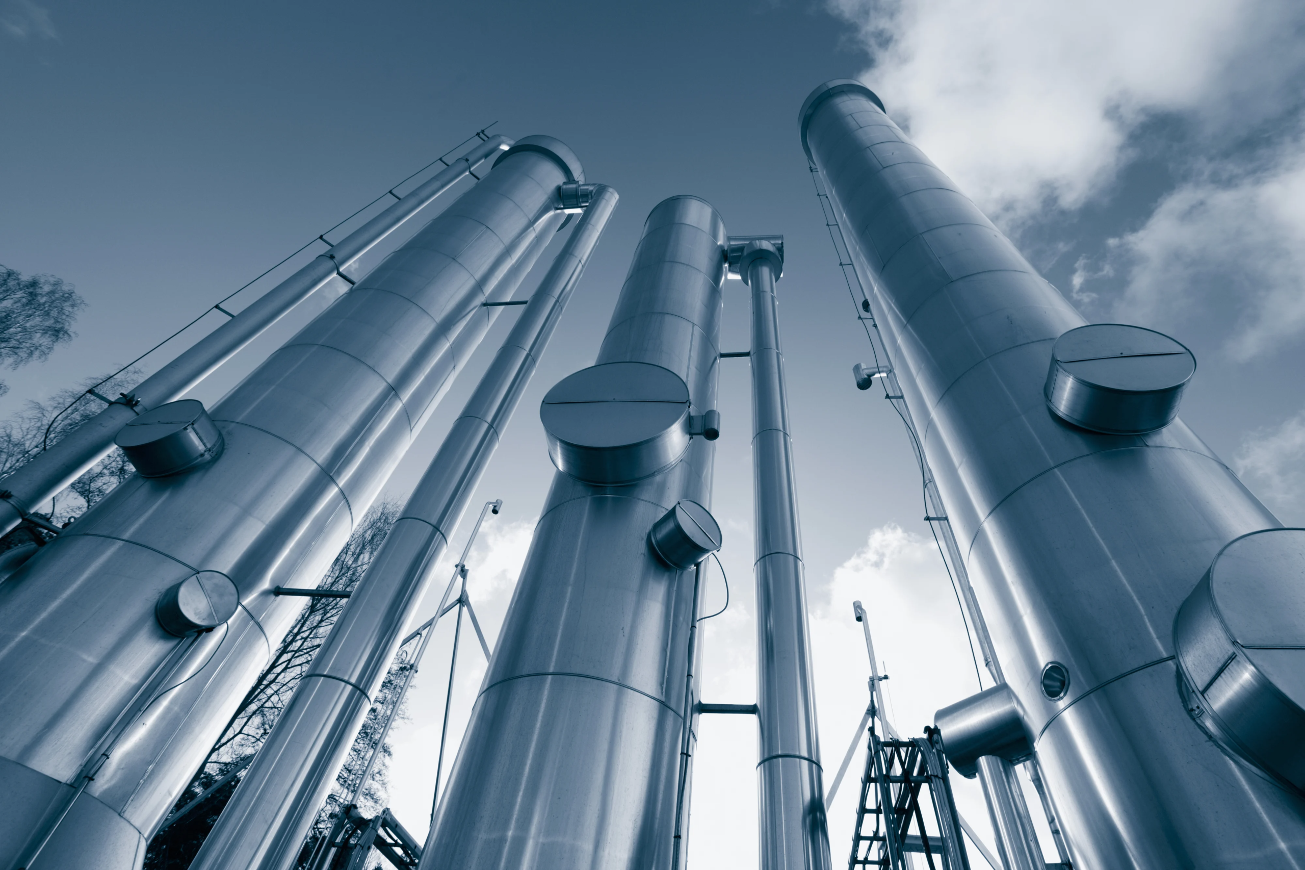 Blue-toned industrial scene looking upward at tall large-diameter stainless steel columns and vessels against cloudy sky